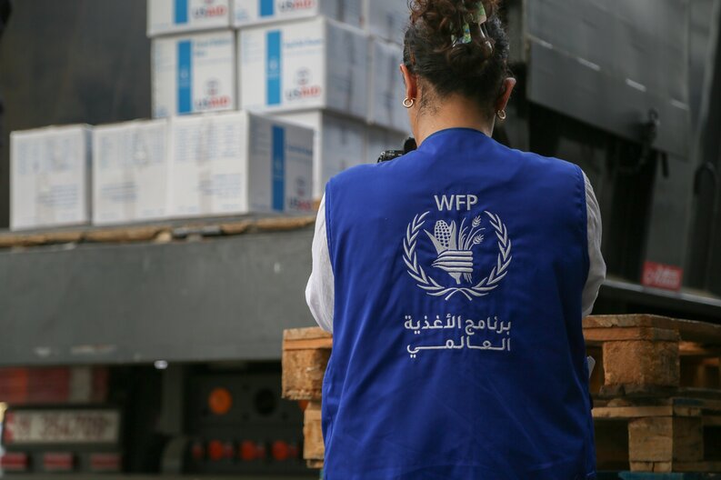 A woman wearing a WFP-vest seen from behind, standing in front of food parcels. © WFP/ Mohammad Awadh