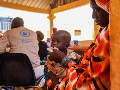 Photo: PAM/Richard Mbouet Un enfant reçoit un soutien nutritionnel dans une clinique du PAM, tandis que des accompagnants et des agents de santé participent à une séance communautaire de sensibilisation à la santé et à la nutrition.