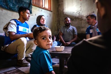 A little boy in Gaza, looking at the camera while his family discusses in the background with WFP workers.