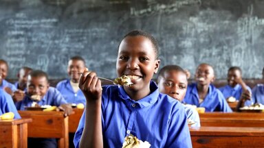 WFP/Aristide Gatera Un enfant à un pupitre d'école avec une fourchette pleine de nourriture