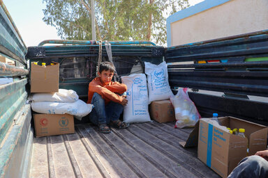 Un garçon vêtu d'une chemise orange et d'un jean bleu est accroupi à l'arrière d'un camion en Syrie, au milieu de sacs et de boîtes contenant de la nourriture du PAM