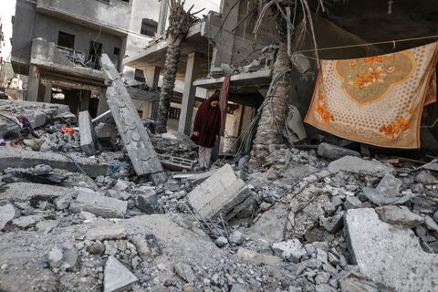 a woman in a black coat and headscarf walks through rubble in Gaza