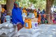 A woman at a distribution centre is sat on white bags as people line up behind
