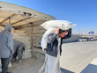 Man carries sack of food on shoulder beside truck loaded with aid bags, while workers stack supplies in a bright open yard under a clear sky.