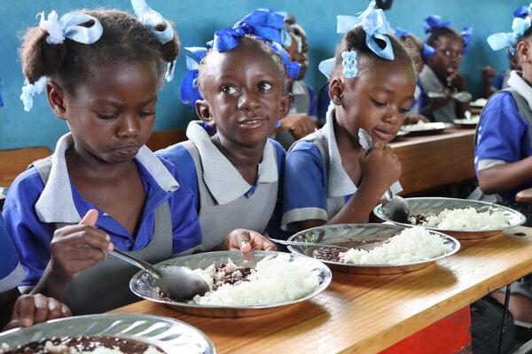 Photo : PAM/Alexis Masciarelli. Des élèves âgés de 5 à 7 ans prennent un repas chaud dans leur classe de l'école primaire Baptiste Salem de Dondon, qui compte 600 élèves. Les repas sont préparés quotidiennement à partir de riz et de haricots achetés à des producteurs locaux, dans le cadre du programme d'alimentation scolaire à base de produits locaux du PAM. 