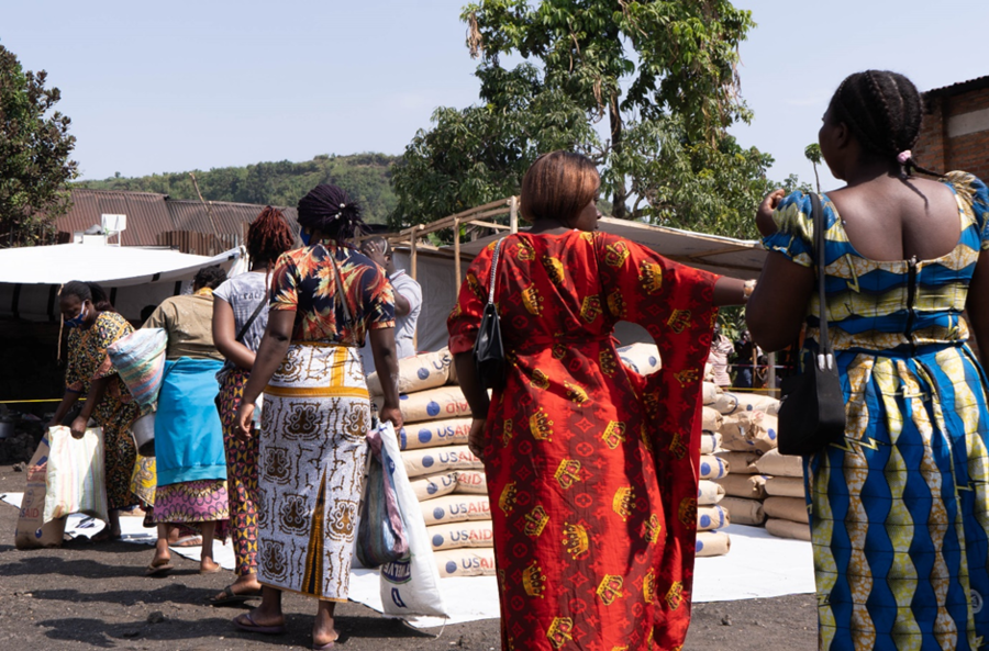 Women line up near stacked bags of food provisions.