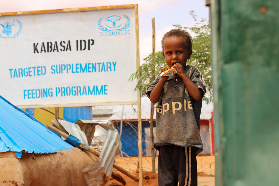 A child eats nutritious food from WFP while standing in front of a sign for a refugee camp