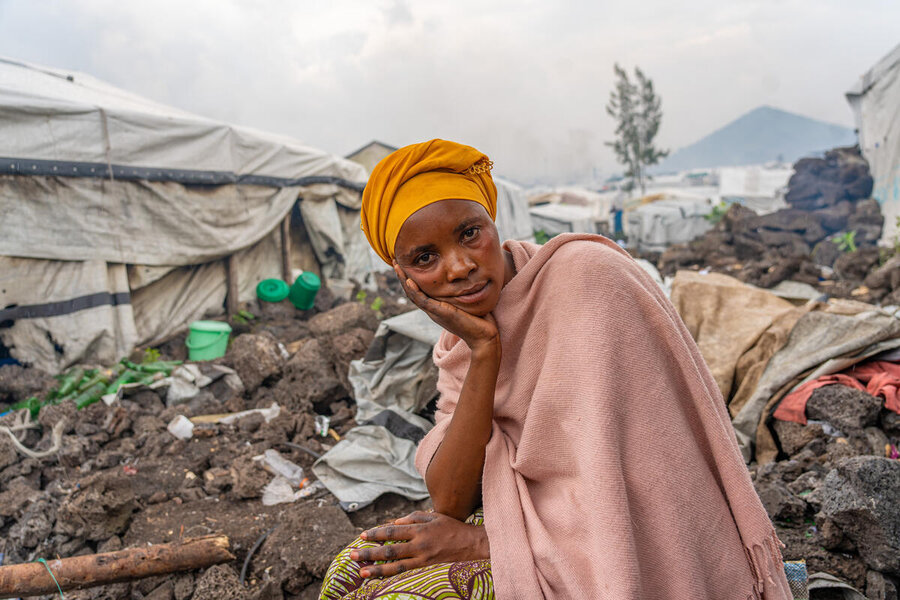 A woman with orange headgear and a pink shawl has her hand to her chin as she sits in front of a row of tarpaulin tents in a displacement camp