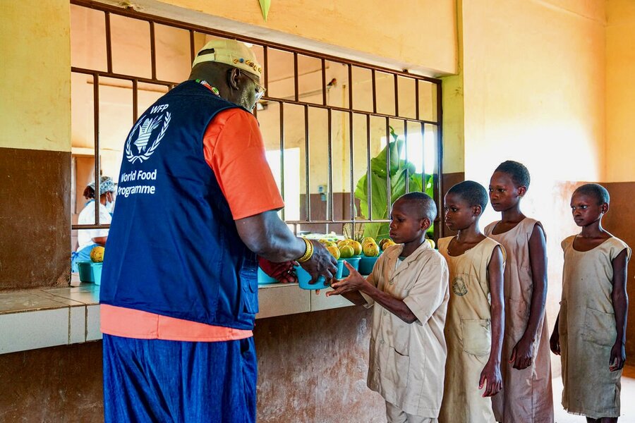 A man sporting blue pants and matching WFP vest serves up blue bowls of food to primary school children in Benin. Photo: WFP/Richard Mbouet