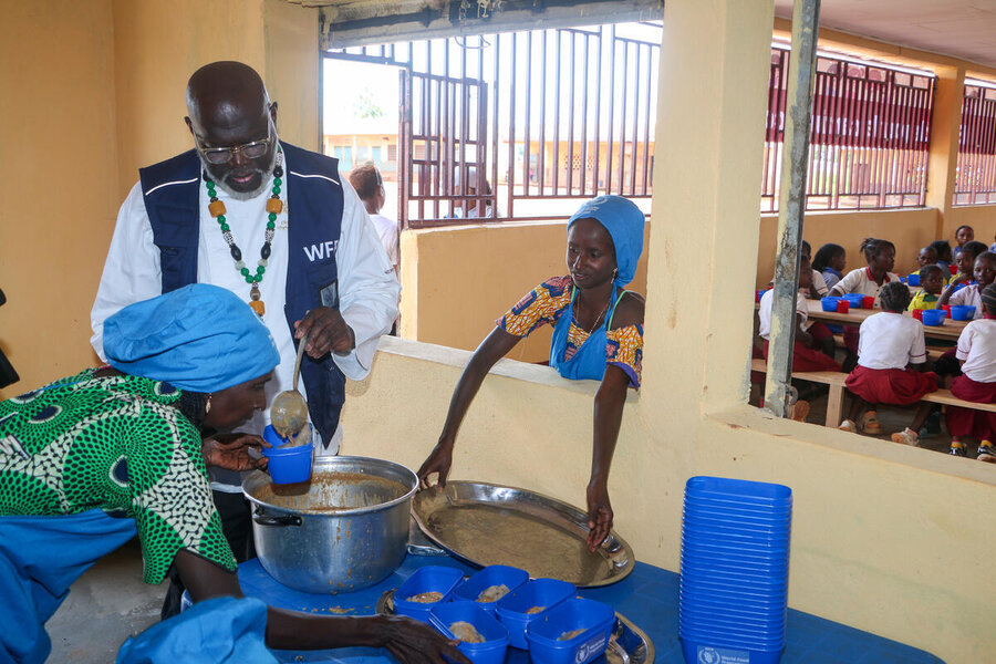 A man wearing a blue WFP vest and African beads helps two female cooks serve up bowlfuls of stew to school children sitting nearby. Photo: WFP/Jordan Onana