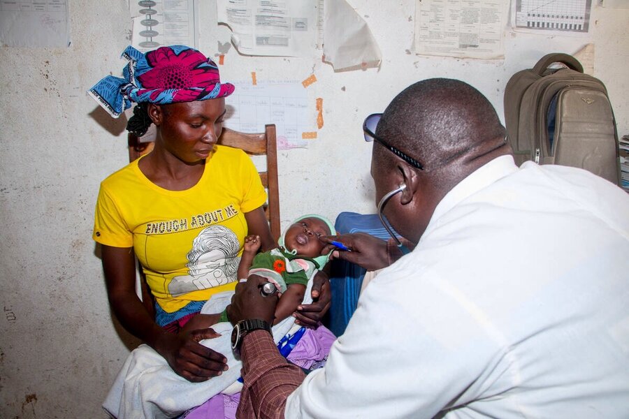 A health worker with a stethoscope checks out a baby held out by her mother. Photo: WFP/Joseph Fambove