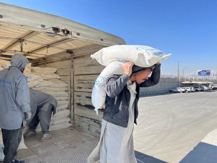 Man carries sack of food on shoulder beside truck loaded with aid bags, while workers stack supplies in a bright open yard under a clear sky.