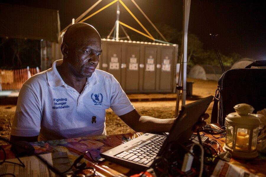 A man with a white WFP T-shift sits outside at night, checking his laptop in this 2015 photo from Guinea, backdropped by a lit-up metal units (an Ebola treatment centre) and a field. Photo: WFP/Rein Skullerud